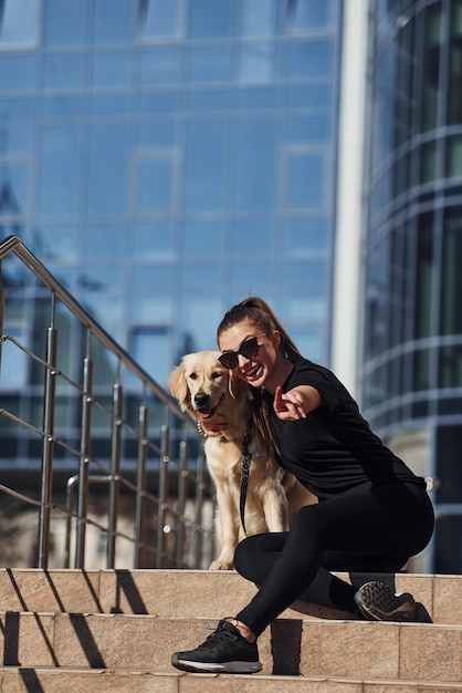 Jonge positieve vrouw die met haar hond op de trap zit als ze buiten wandelt in de buurt van een bedrijfsgebouw