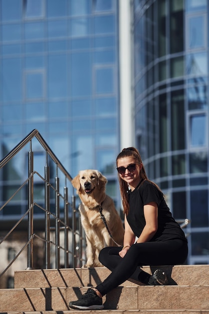 Jonge positieve vrouw die met haar hond op de trap zit als ze buiten wandelt in de buurt van een bedrijfsgebouw