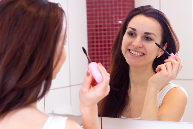 Jonge positieve vrouw die roze mascara gebruikt voor de spiegel in haar bordeauxrode badkamer