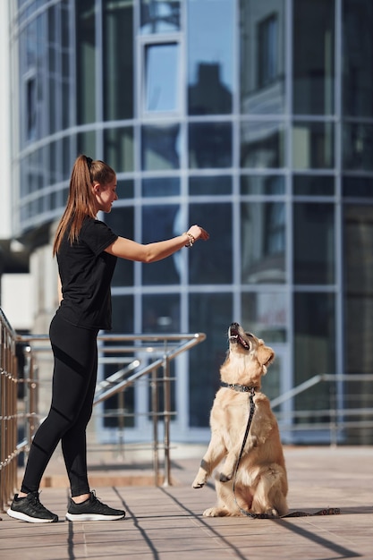 Jonge positieve vrouw heeft plezier en doet trucjes met haar hond wanneer ze buiten in de buurt van een bedrijfsgebouw gaat wandelen