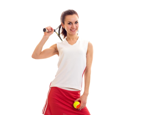 Jonge positieve vrouw in wit sportshirt en rode rok met tennisracket en gele bal