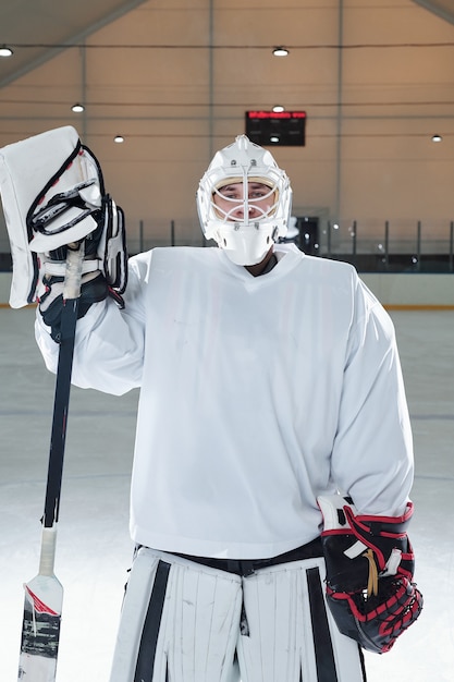 Jonge professionele hockeyspeler in sportuniform, handschoenen en beschermende helm die stok vasthoudt terwijl hij na het spelen op de ijsbaan voor de camera staat