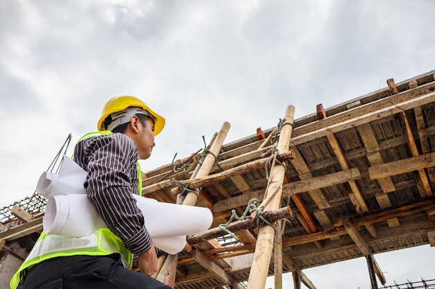 Jonge professionele ingenieur werknemer in beschermende helm en blauwdrukken papier bij de hand bezig met ladder op de bouwplaats van het huis