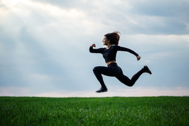 Foto jonge professionele turnster springt in de natuur tegen de blauwe lucht. meisjesatleet in een zwarte top en zwarte legging doet acrobatische oefeningen