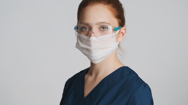 Jonge professionele vrouwelijke arts met een beschermende bril en een medisch masker die aandachtig in de camera kijkt, geïsoleerd Safety first concept