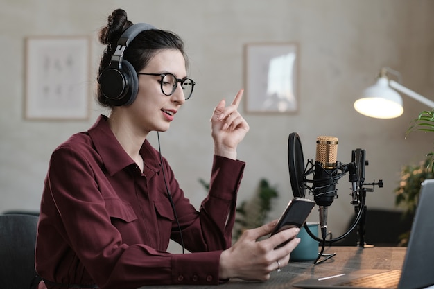 Jonge radio-dj in koptelefoon die online praat met haar mobiele telefoon aan de tafel in de studio