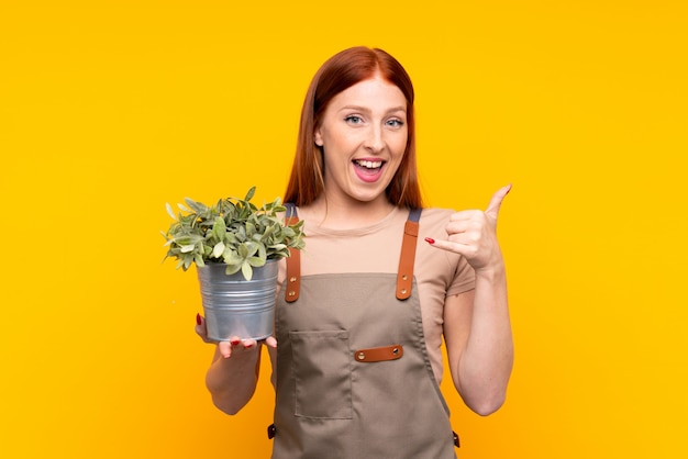 Jonge redhead tuinmanvrouw die een installatie over geïsoleerde gele muur houden die telefoongebaar maken