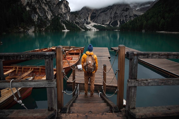 Jonge reiziger jongen met blauwe hoed, vintage rugzak en gele regenjas op steiger van Lago di Braies omgeven door bergen van Italiaanse Alpen