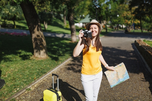 Jonge reiziger toeristische vrouw in hoed met koffer, stadsplattegrond foto's maken op retro vintage fotocamera in de stad buiten. Meisje dat naar het buitenland reist om een weekendje weg te reizen. Toeristische reis levensstijl.