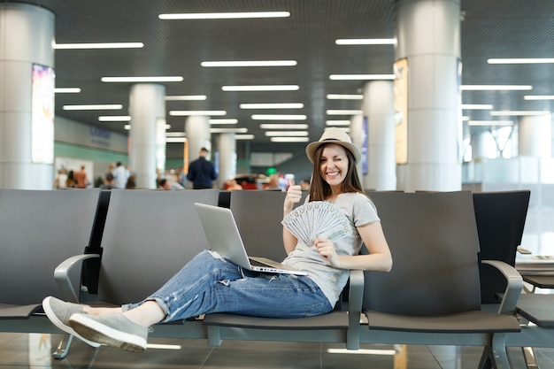 Jonge reiziger toeristische vrouw met laptop, bundel dollars vasthouden, contant geld duim omhoog knipperend wachten in lobby hal op luchthaven in
