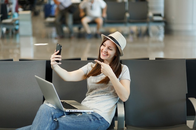 Jonge reizigerstoeristenvrouw die aan laptop werkt die selfie op mobiele telefoon doet die met de vinger op de camera wijst, wacht in de lobbyhal op de luchthaven