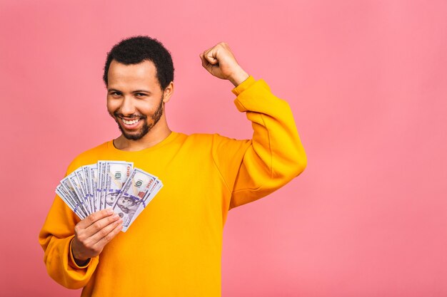 Jonge rijke gelukkig man in casual holdingsgeld dollarbiljetten met verrassing geïsoleerd over roze muur