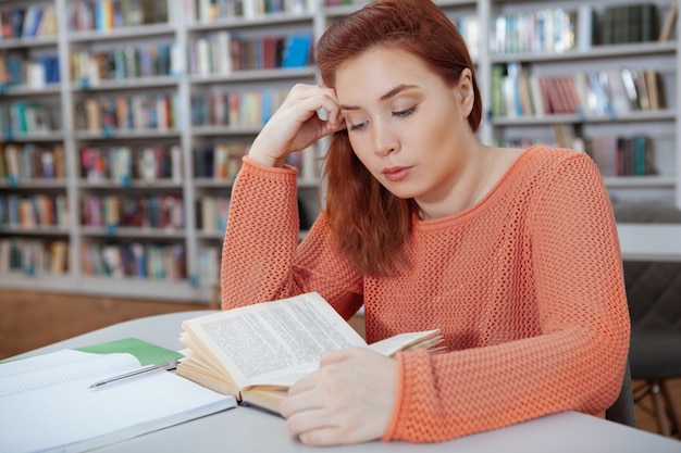 Jonge rode haired vrouw die een boek leest bij lokale bibliotheek