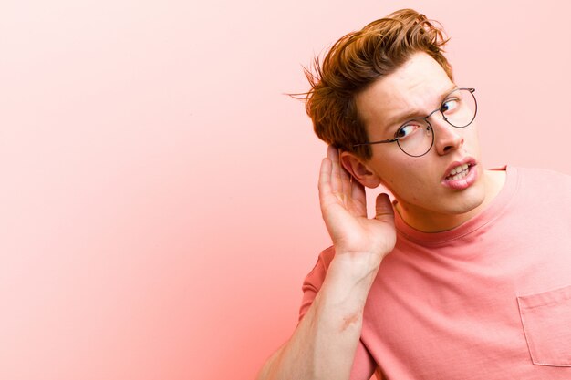 Jonge rode hoofdmens die ernstig en nieuwsgierig kijkt, luistert, probeert een geheim gesprek of roddel te horen, afluisterend over roze muur