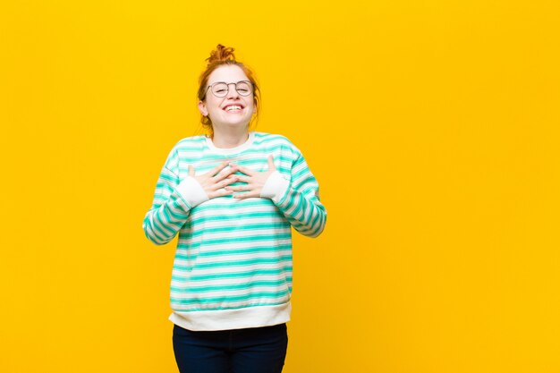 Jonge rode hoofdvrouw die gelukkig, verrast, trots en opgewekt kijkt, richtend aan zich tegen oranje muur