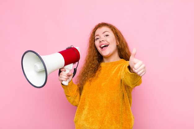 Jonge rode hoofdvrouw die trots, zorgeloos, zelfverzekerd en gelukkig voelt, positief glimlachend met duimen omhoog met een megafoon