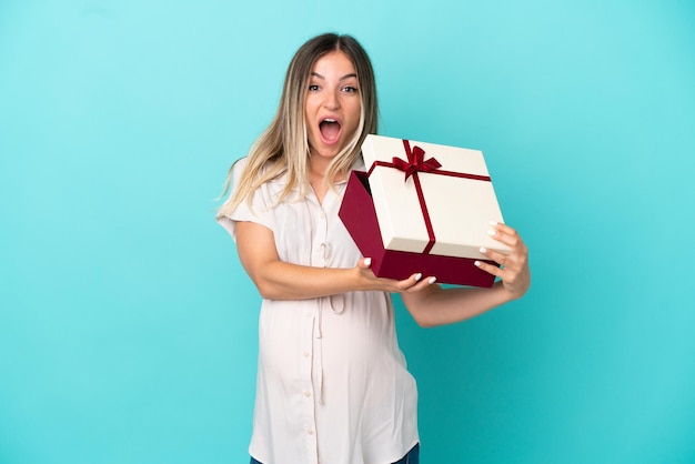 Jonge Roemeense vrouw geïsoleerd op blauwe achtergrond zwanger en met een geschenk met verbaasde uitdrukking