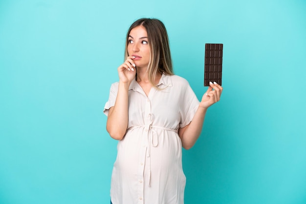 Jonge Roemeense vrouw geïsoleerd op blauwe achtergrond zwanger en twijfels terwijl ze chocolade vasthoudt