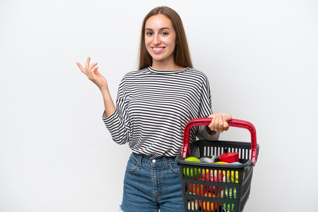 Jonge Roemeense vrouw met een winkelmandje vol eten geïsoleerd op een witte achtergrond handen uitstrekkend naar de zijkant om uit te nodigen om te komen