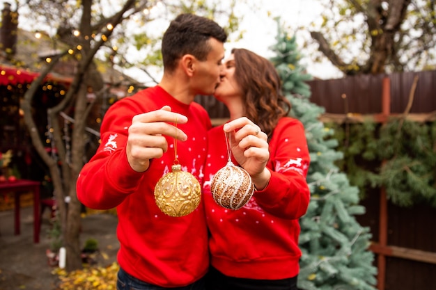 Foto jonge romantische paar ballen op de boom houden en zoenen op de achtergrond. ze versieren de kerstboom buiten voor kerstmis.