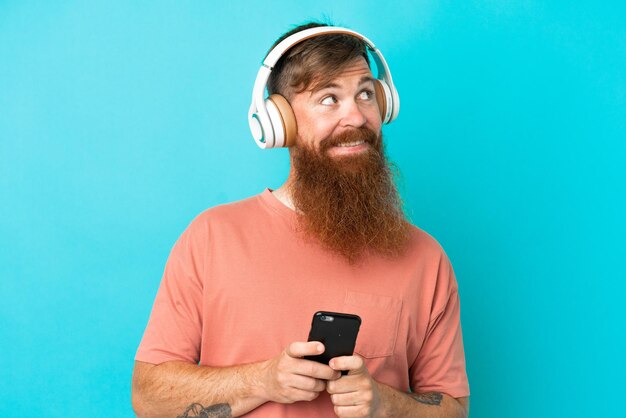 Jonge roodachtige blanke man geïsoleerd op blauwe achtergrond muziek luisteren met een mobiel en denken