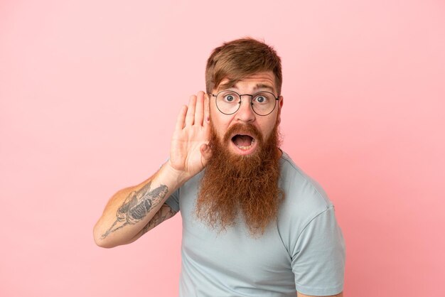 Foto jonge roodachtige blanke man geïsoleerd op roze achtergrond luisteren naar iets door hand op het oor te leggen