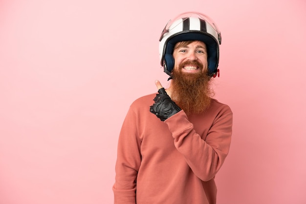 Jonge roodachtige blanke man met een motorhelm geïsoleerd op een roze achtergrond die naar de zijkant wijst om een product te presenteren