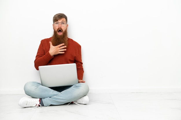 Jonge roodachtige blanke man met laptop geïsoleerd op een witte achtergrond verrast en geschokt terwijl hij naar rechts kijkt