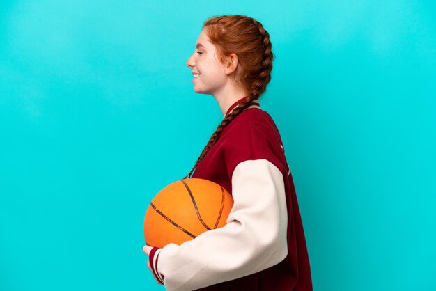 Jonge roodachtige vrouw die basketbal speelt geïsoleerd op een blauwe achtergrond die lacht in zijpositie