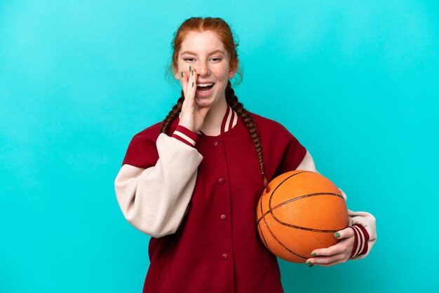 Jonge roodachtige vrouw die basketbal speelt geïsoleerd op een blauwe achtergrond die met wijd open mond schreeuwt