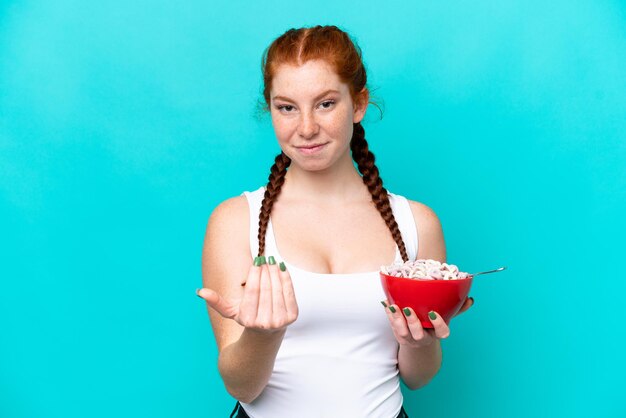 Jonge roodachtige vrouw met een kom ontbijtgranen geïsoleerd op een blauwe achtergrond die uitnodigt om met de hand te komen Blij dat je bent gekomen