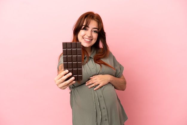 Jonge roodharige blanke vrouw geïsoleerd op roze achtergrond zwanger en met chocolade