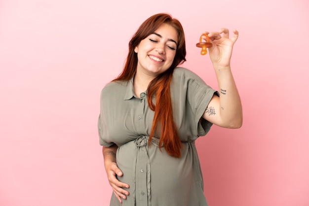 Jonge roodharige blanke vrouw geïsoleerd op roze achtergrond zwanger en met een fopspeen