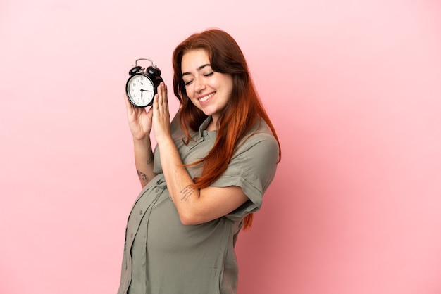 Jonge roodharige blanke vrouw geïsoleerd op roze achtergrond zwanger en met klok