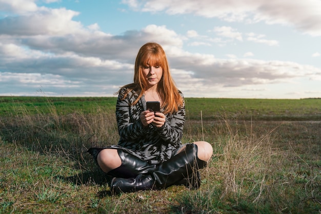 Jonge roodharige blanke vrouw zittend op het gras in een veld met haar telefoon gekleed in cowboylaarzen onder een met wolken bedekte hemel