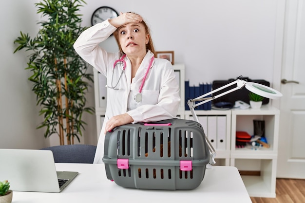 Jonge roodharige dierenarts vrouw die werkt bij dierenkliniek gestrest en gefrustreerd met hand op hoofd, verrast en boos gezicht