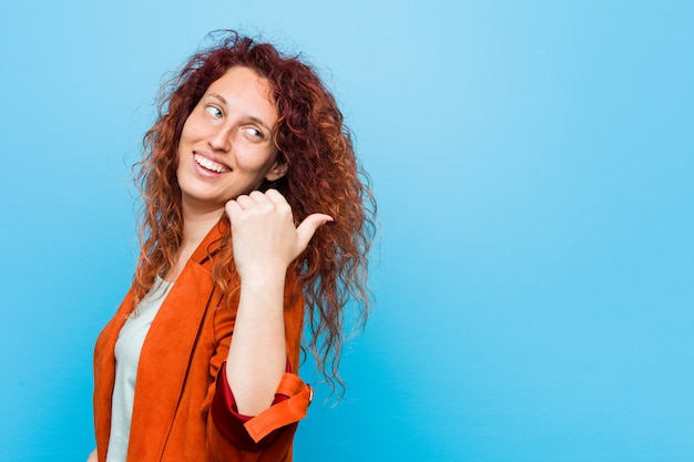 Jonge roodharige elegante vrouw wijst met duimvinger weg, lachend en zorgeloos.