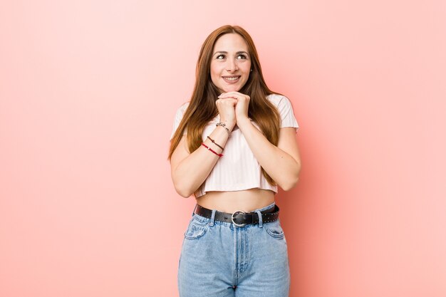 Jonge roodharige gembervrouw tegen een roze muur houdt de handen onder de kin, kijkt vrolijk opzij.