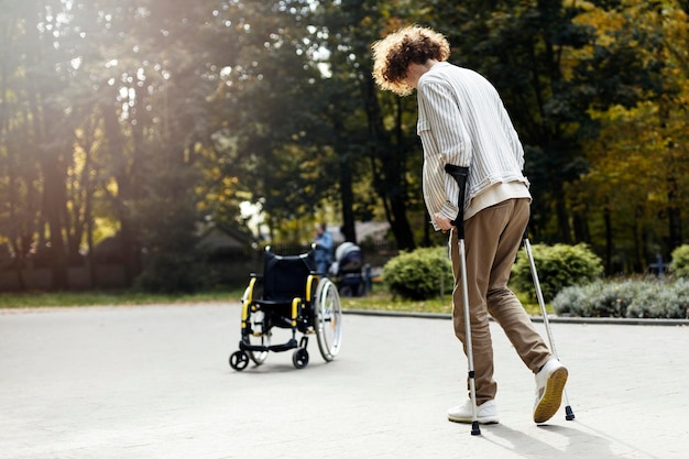 Jonge roodharige jongen op krukken loopt naar een rolstoel op straat Man in casual kleding achteraanzicht