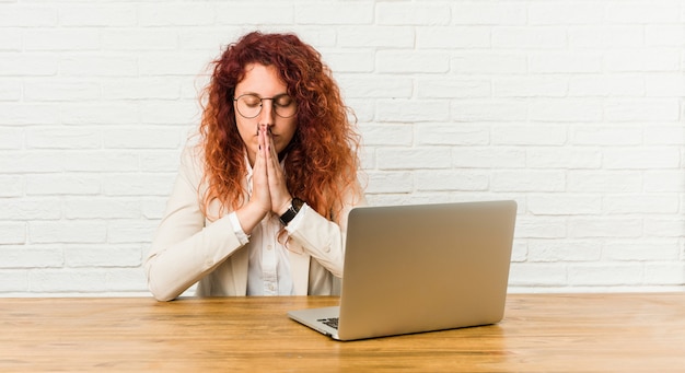 Jonge roodharige krullende vrouw die met haar laptop hand in hand indient bidt in de buurt van mond, voelt zich zeker.