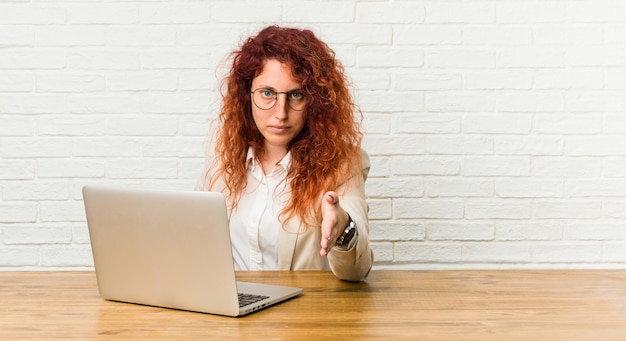 Jonge roodharige krullende vrouw die met haar laptop uitrekkende hand werkt bij camera in groetgebaar.