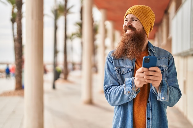 Jonge roodharige man die zelfverzekerd glimlacht met zijn smartphone op straat