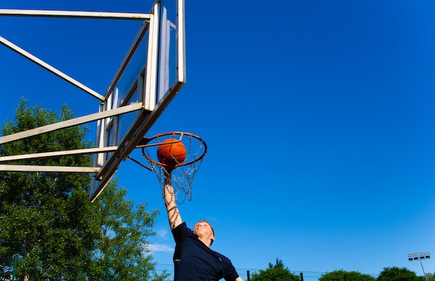 Jonge roodharige man in een donkerblauw t-shirt gooit een bal in beweging in een basketbalring tegen een blauwe lucht buiten