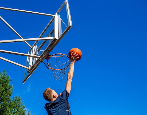 Jonge roodharige man in een donkerblauw t-shirt gooit een bal in beweging in een basketbalring tegen een blauwe lucht buiten