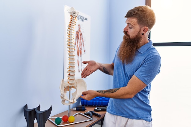 Jonge roodharige man met fysiotherapeut uniform wijzend naar anatomisch model van de wervelkolom in de kliniek voor fysiotherapie