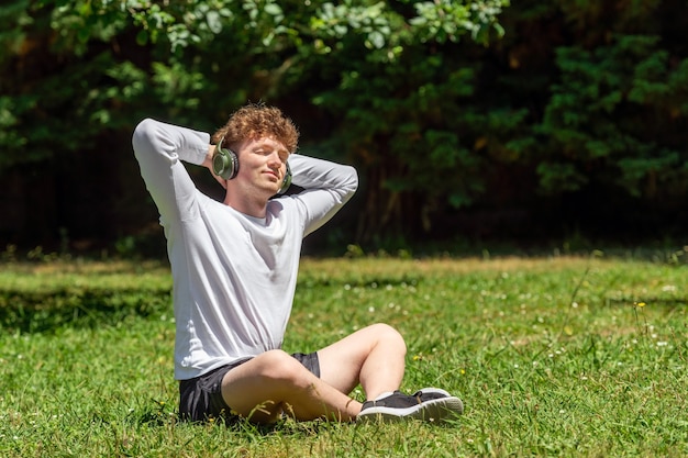 Jonge roodharige man met koptelefoon zittend op het groene gras op een zonnige dag