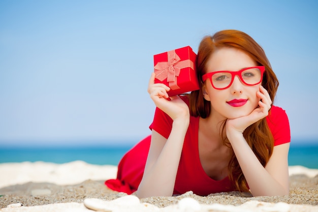 Jonge roodharige meisje in een rode jurk met geschenkdoos rust op zomer zee strand