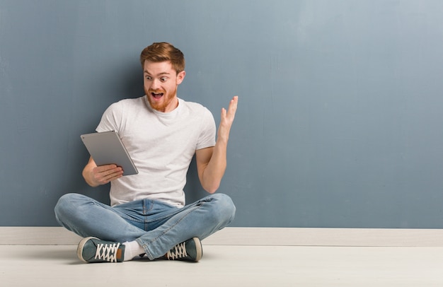 Jonge roodharige student man zittend op de vloer een overwinning of succes te vieren. hij houdt een tablet vast.