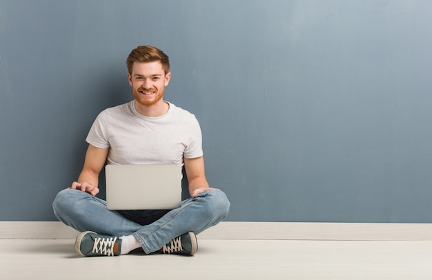 Jonge roodharige student man zittend op de vloer vrolijk met een grote glimlach Hij houdt een laptop