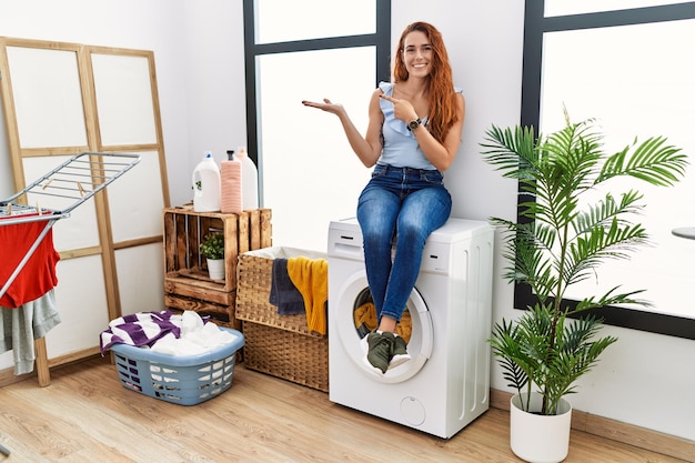 Jonge roodharige vrouw die de was doet zittend op de wasmachine verbaasd en glimlachend naar de camera terwijl ze met de hand presenteert en met de vinger wijst